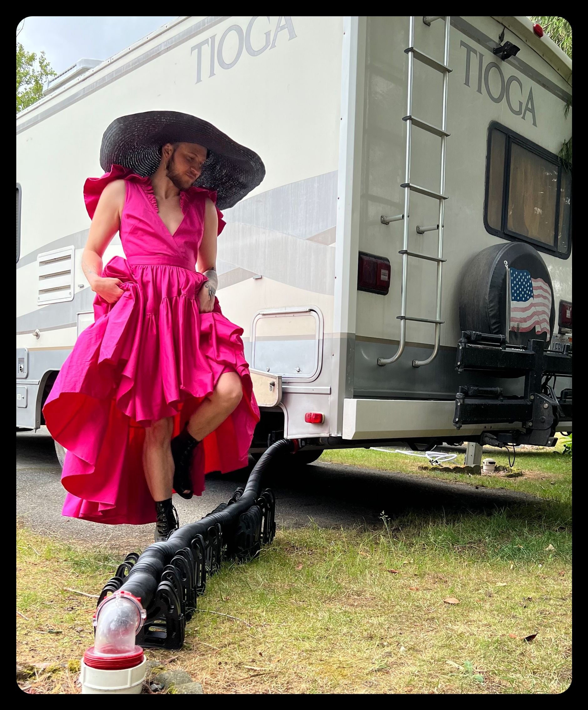 Photo: A man wearing a magenta dress with tiered ruffles and an absurdly wide black straw hat steps over a sewage hose connected to an RV at one end and a dump portal in the ground at the other.