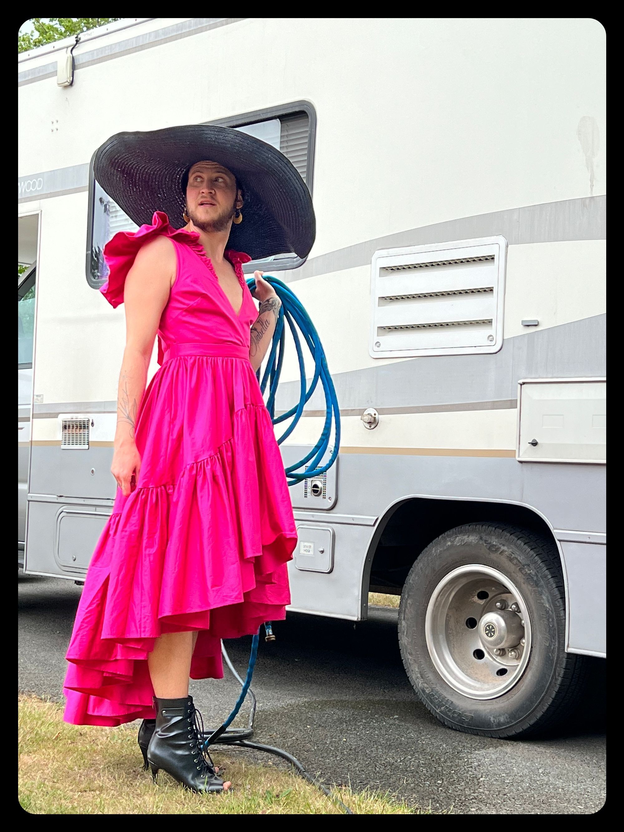 Photo: A man in a magenta ruffled dress and oversize black straw hat holds a hose outside an RV.