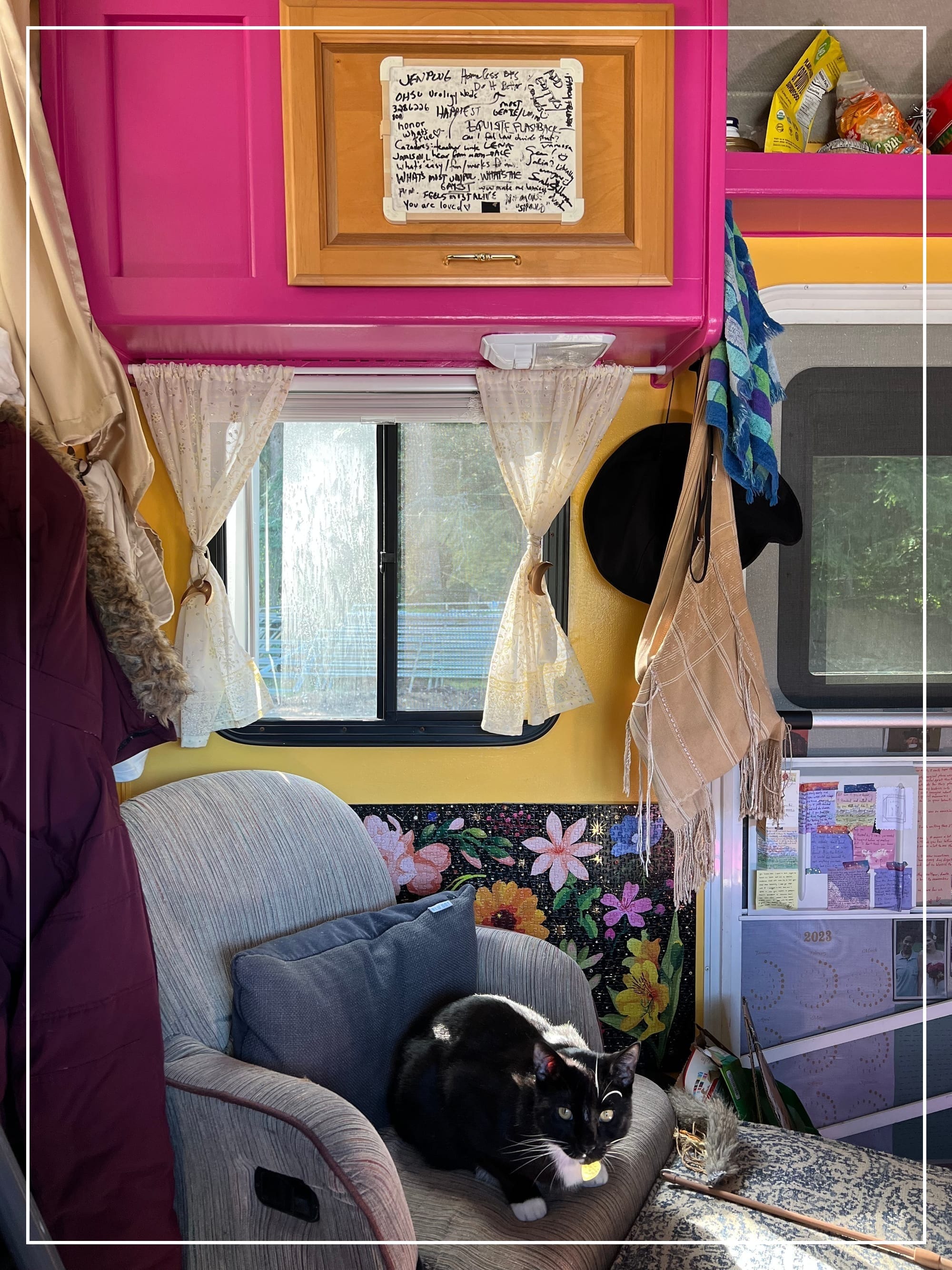 A black cat sits on a chair in a sunny, brightly colored RV. The cupboard above has a dry-erase board crowded with scrawled writing.