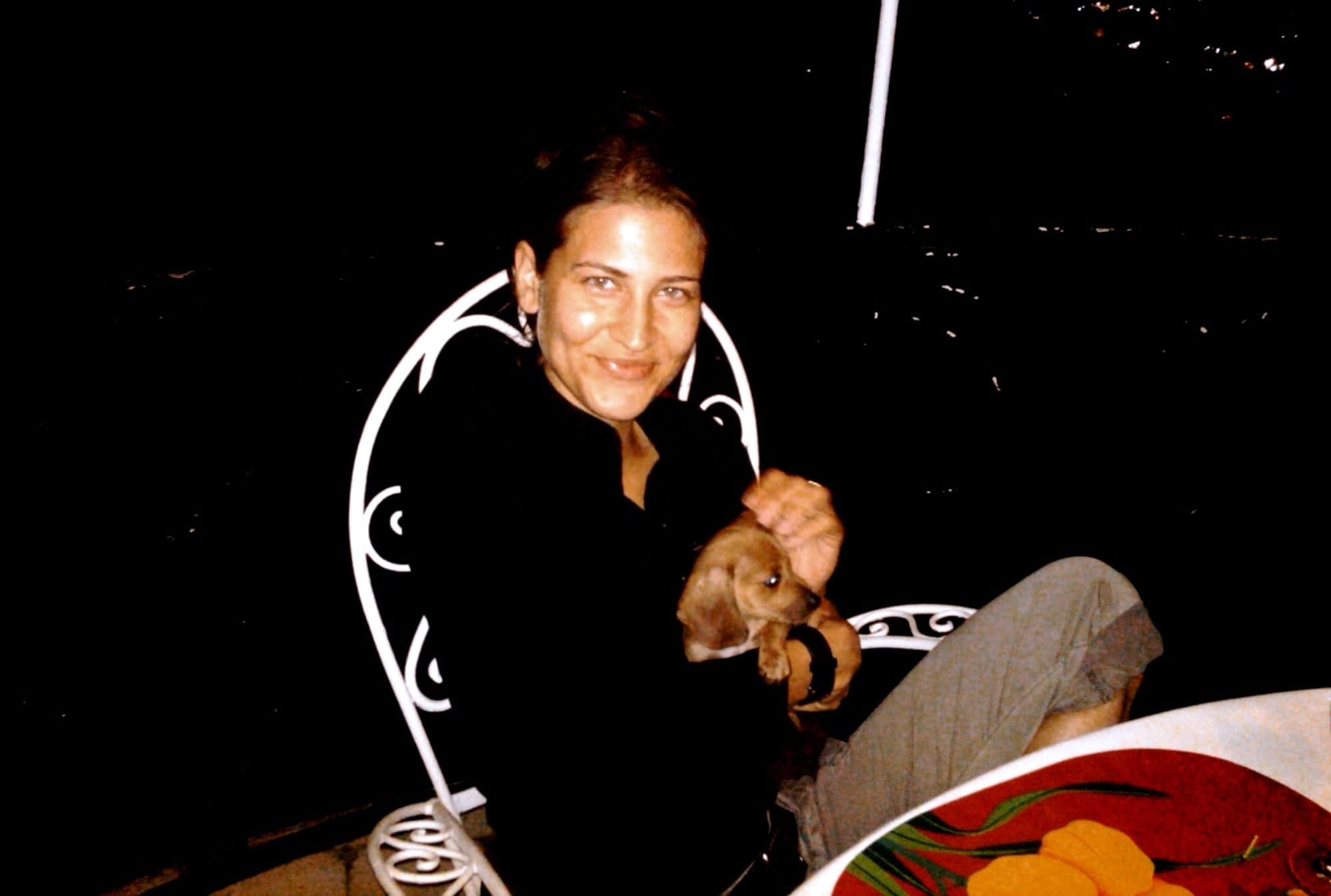 A person in a black long-sleeve shirt holds a small brown puppy with floppy ears. It is very dark in the background, and they sit at a table with white tablecloth on a balcony.