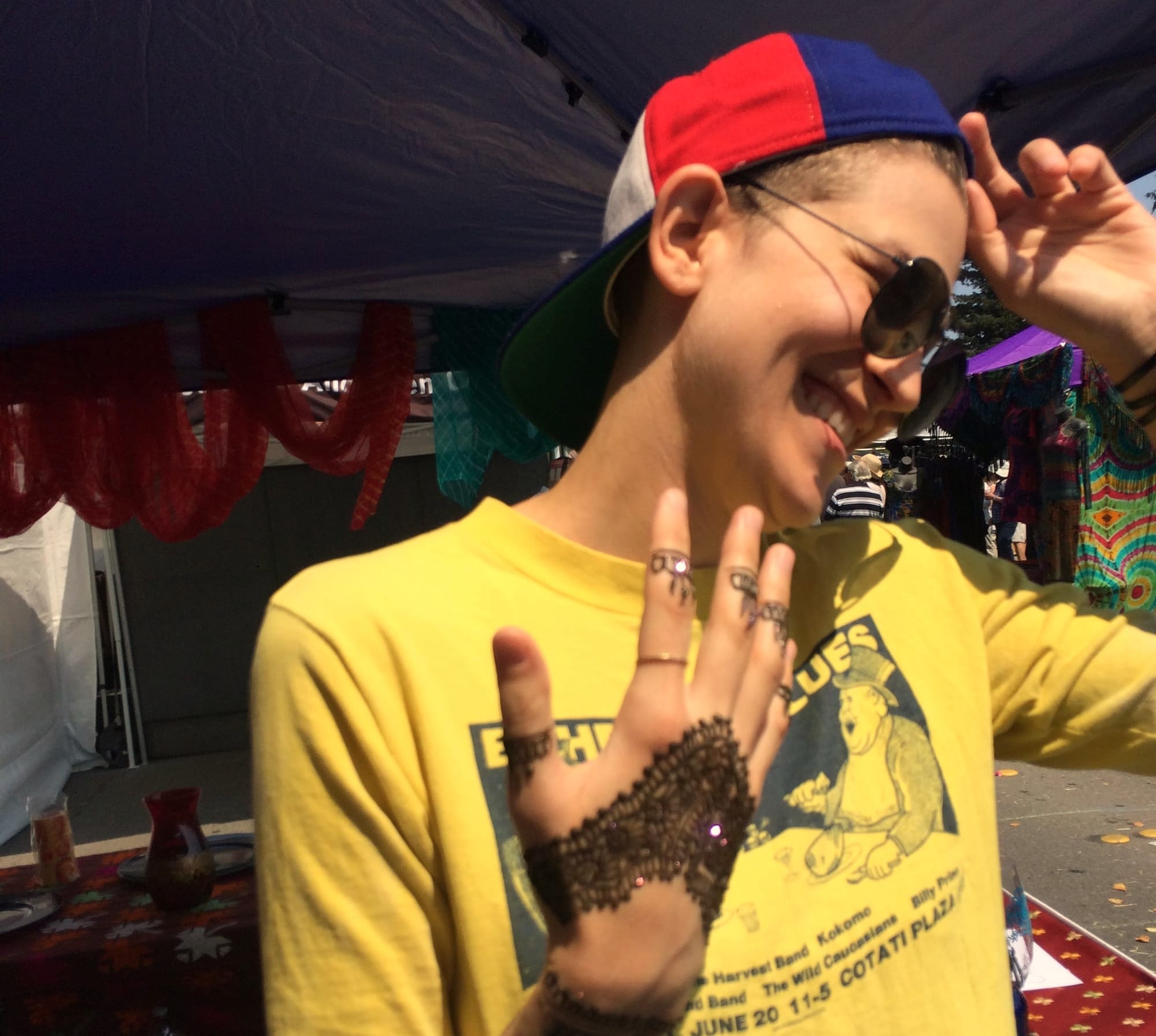 A person with short hair and breasts in a yellow t-shirt, ball cap, and black-hennaed hands.