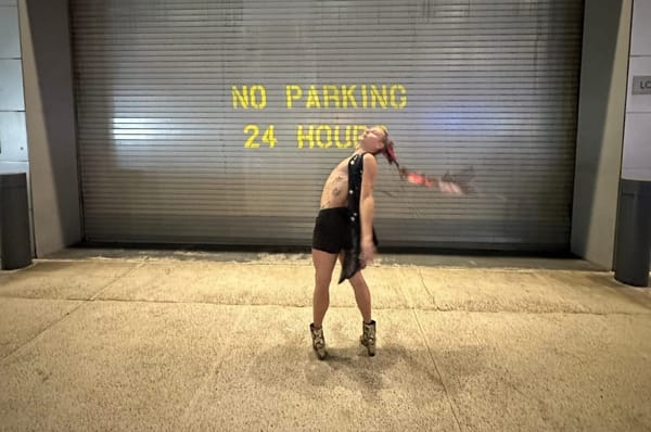 A person in high-heeled alligator boots, shorts, and a sequin vest stands in front of a garage painted NO PARKING.