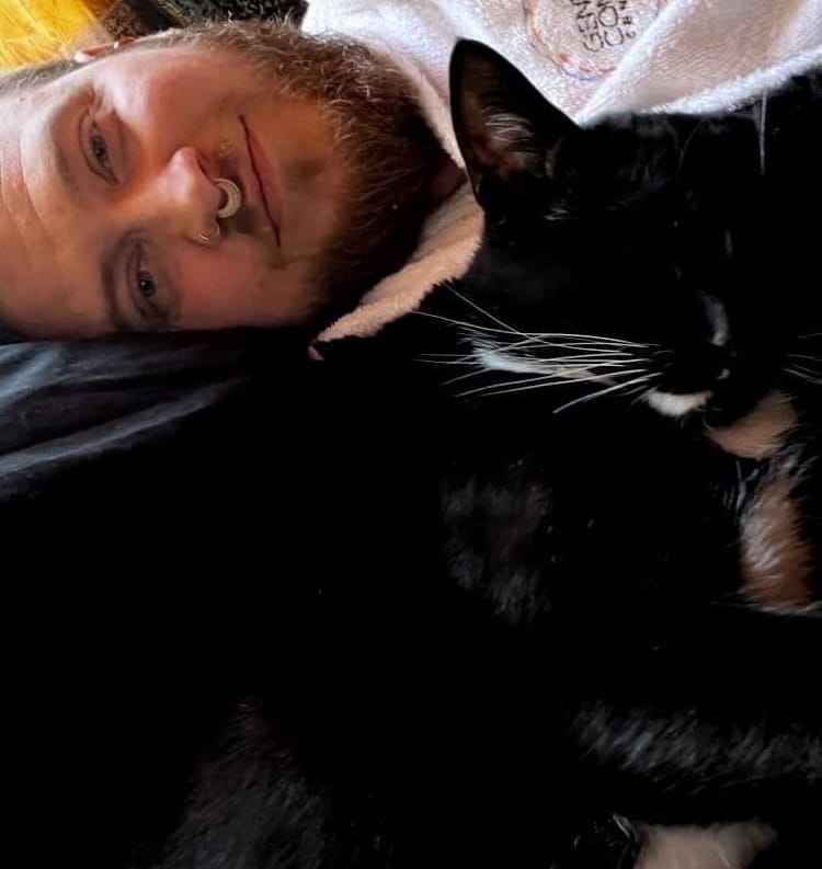 A man with a beard lying next to a sleeping black cat with white whiskers.