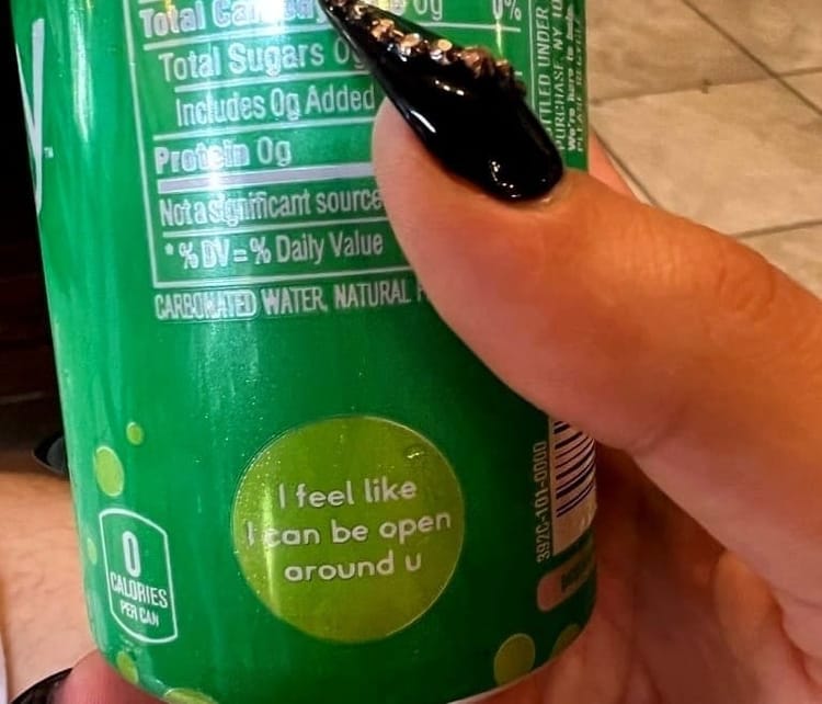 A photograph of a sparkling water can reading “I feel like I can be open around u,” held by long painted nails.