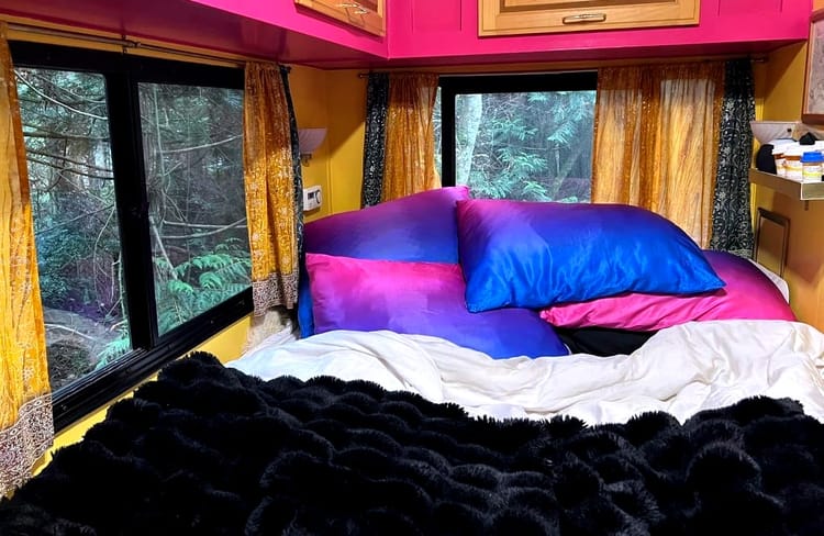 Four pillows encased in ombre silk fading from royal blue to pink on an RV bed in front of windows overlooking green trees.