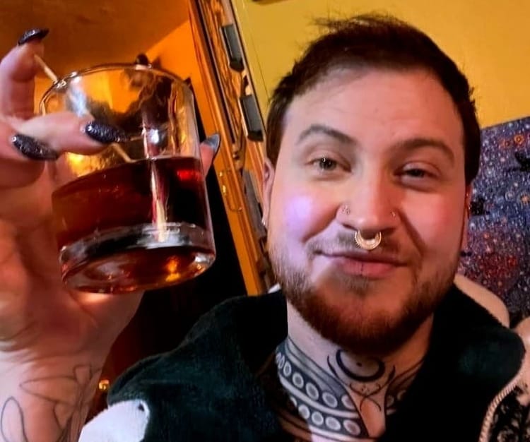 A man with sparkling black stiletto nails holds up a glass of whiskey in a “cheers” motion.