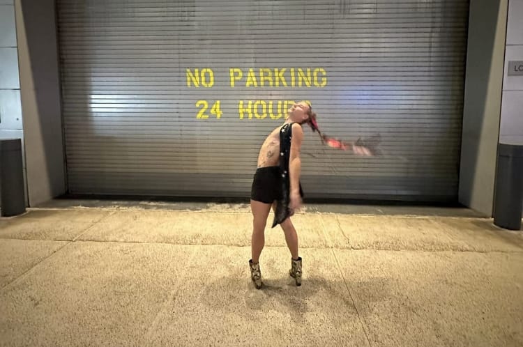 A person in high-heeled alligator boots, shorts, and a sequin vest stands in front of a garage painted NO PARKING.