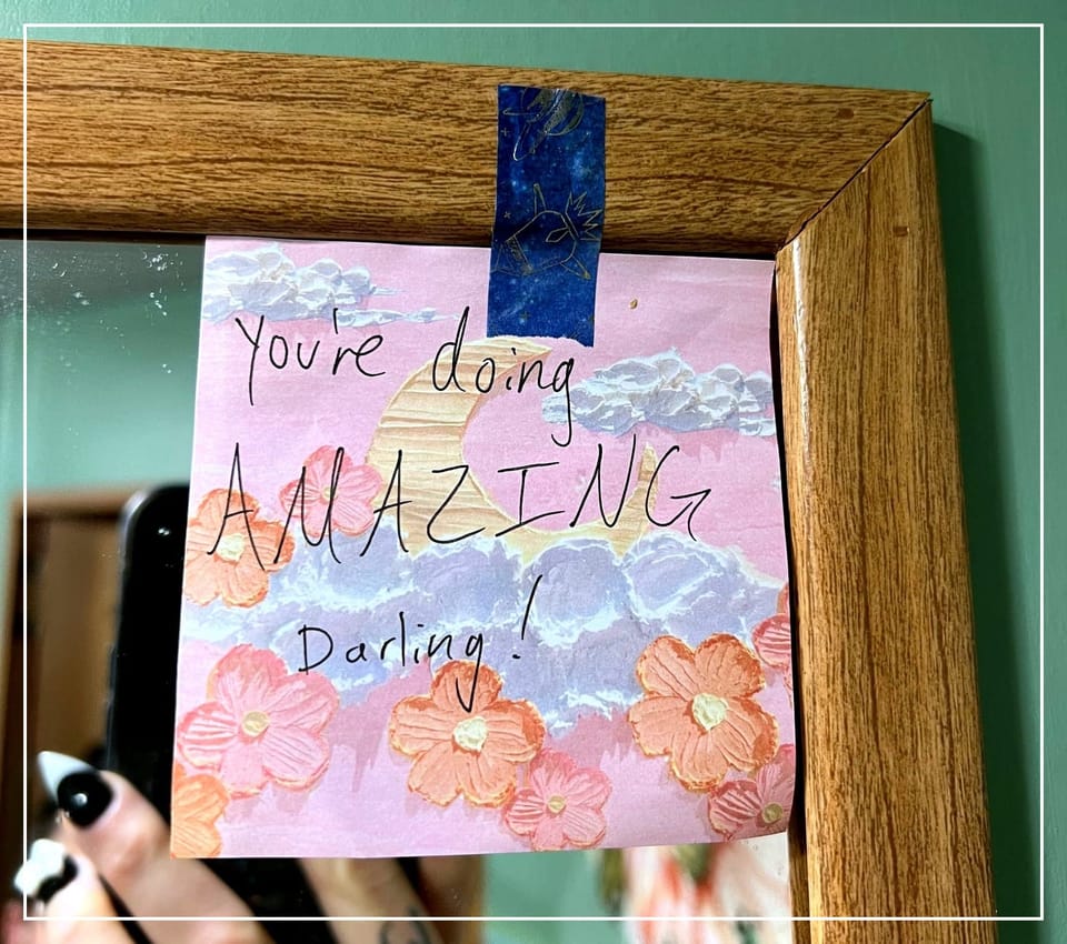 A small blue and pink pastel note taped to a mirror that says: “You’re doing AMAZING darling!”