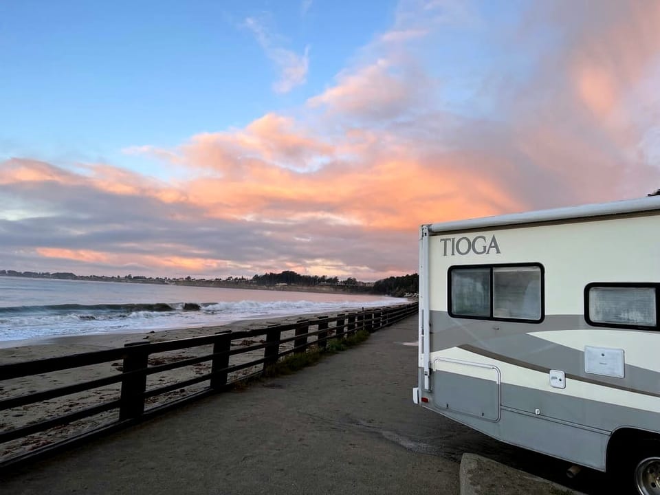 The back end of an RV parked close to a beach with low waves washing on it. Overhead the blue sky is streaked with clouds.