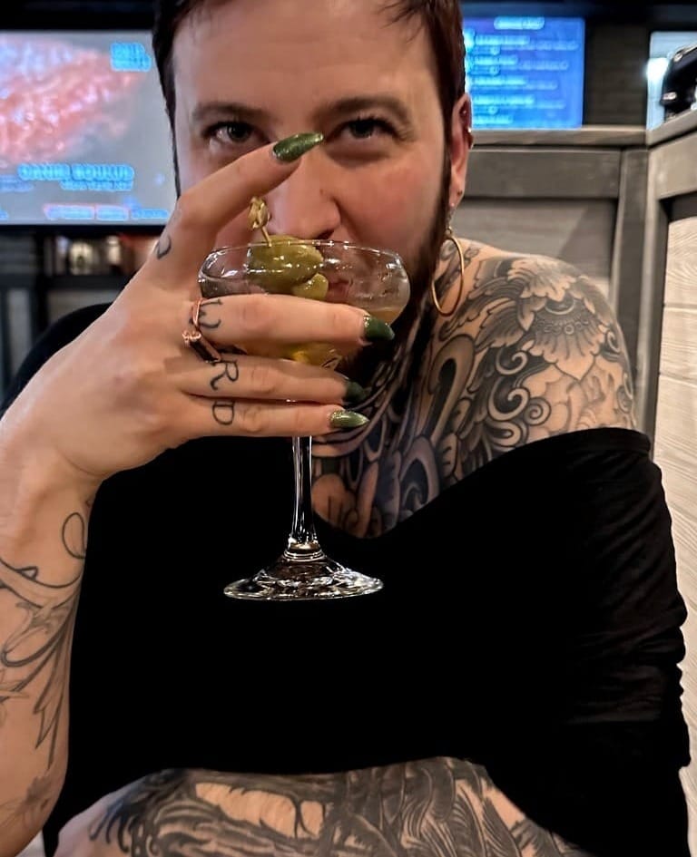 A man with long green nails and covered in black tattoos sips from a martini glass in a restaurant.