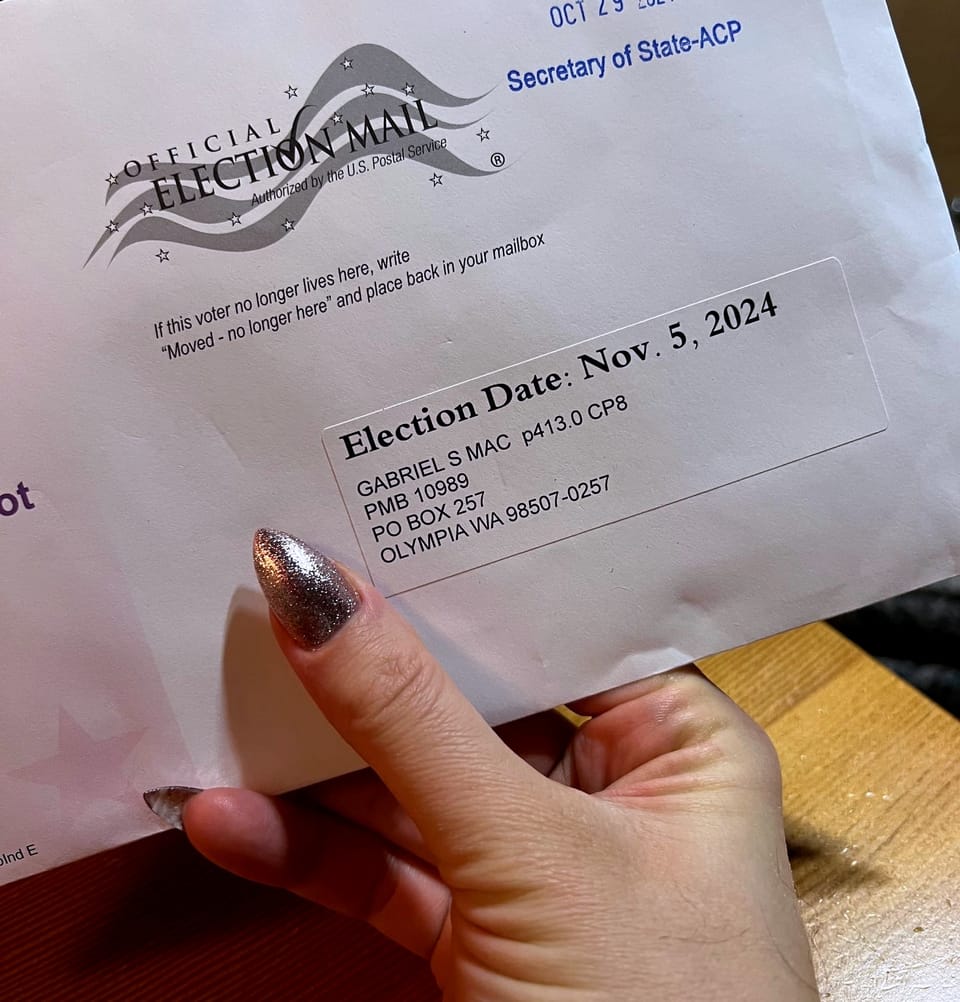 A hand with long gold nails holding a mail-in election ballot addressed to a PO Box in Olympia, Washington.